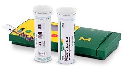 Two cylindrical containers of diagnostic test strips positioned next to a green box labeled with a yellow and green design and medical symbols. The containers have labels providing information about the test strips inside, and one container is open with a strip partially extended, suggesting readiness for use in a clinical or laboratory setting.
