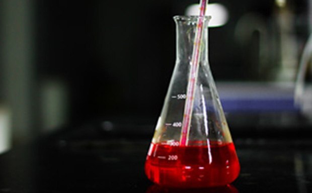 A red liquid is being poured into a graduated Erlenmeyer flask, marked at 200, 300, and 500 milliliters, on a dark laboratory countertop.