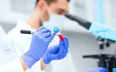 A person in a white lab coat stands in a laboratory facing away from the camera, holding a clipboard and writing notes. The laboratory is equipped with shelves stocked with various lab equipment such as beakers, flasks, test tubes, and bottles containing chemicals labeled with different colors. A person in a white lab coat stands in a laboratory facing away from the camera, holding a clipboard and writing notes. The laboratory is equipped with shelves stocked with various lab equipment such as beakers, flasks, test tubes, and bottles containing chemicals labeled with different colors.