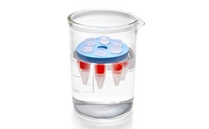 A clear laboratory beaker filled with colorless liquid, containing a small cylindrical container with a red liquid inside. The container is topped with a blue lid, floating on the surface of the liquid in the beaker. The beaker has measurement markings on the side, indicating its volume.