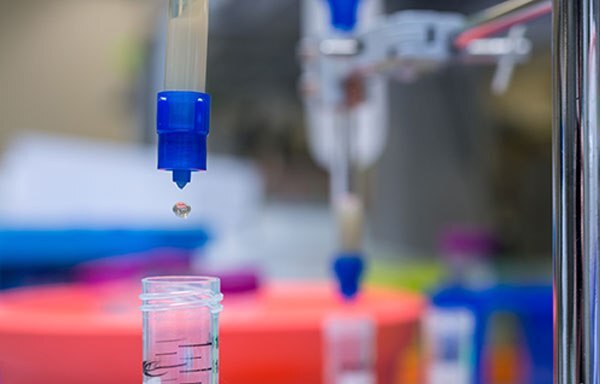 The image captures a precise moment in a laboratory setting, where a blue pipette is dispensing a single clear liquid droplet into a small clear container. 