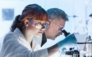 Two people in a lab, wearing white lab coats. One person is looking into a microscope.
