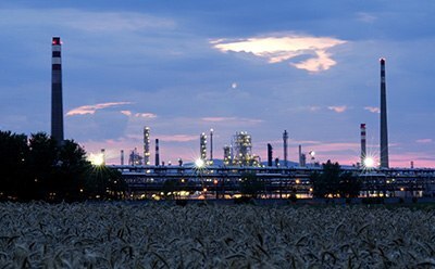 industrial area - petroleum refinery An industrial complex with multiple chimneys and structures illuminated against the twilight sky, with a foreground of darkened crops.