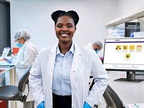 Scientist in a white lab coat and blue gloves standing in a bright laboratory; colleagues work at benches; a desktop monitor shows app icons.