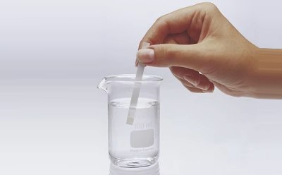 A hand is holding a test strip above a beaker filled with clear liquid. The beaker is labeled with a measurement of 100 ml and is placed on a light background. The test strip is being lowered into the water.